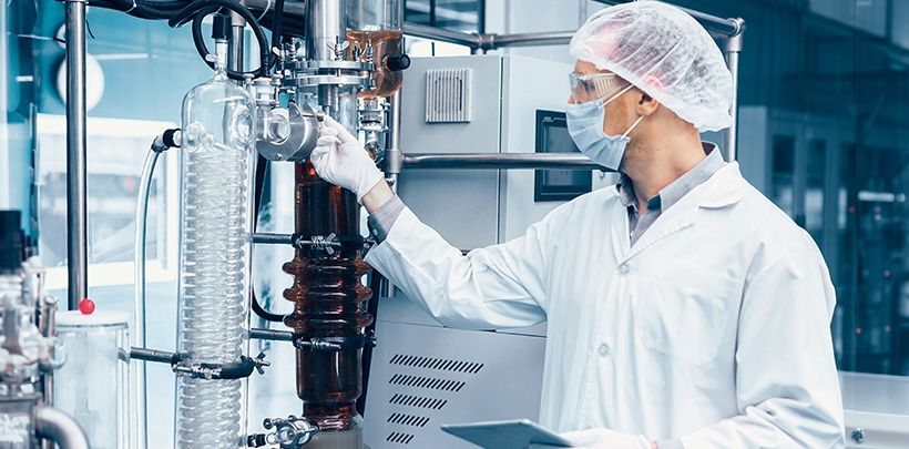 Ein Chemiker arbeitet im Labor von Hänseler an einer Maschine. Er trägt Munschutz, Haarnetz und Brille und hat ein Tablet in der Hand.