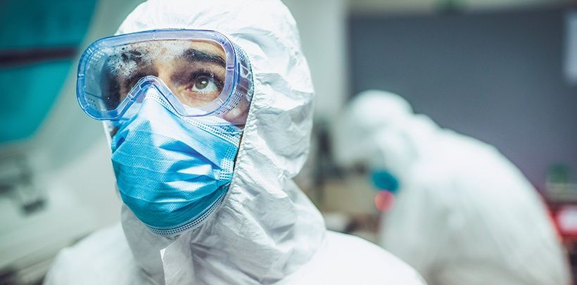 Foto eines Chemikers, der eine Schutzbrille, Mundschutz und Schutzanzug trägt und nach oben schaut. Im Hintergrund ist ein Kollege unscharf zu sehen.